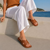 Miele | Close-up of model wearing tan brown leather flat sandals against a coastal beach background