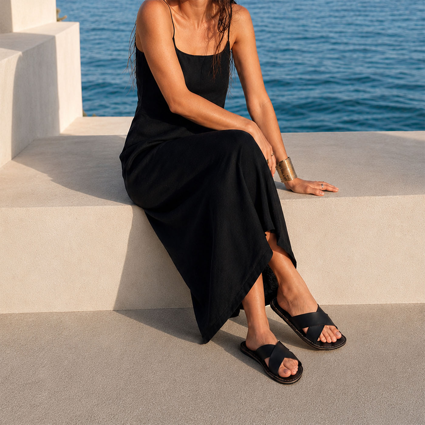 Nero | Model wearing black leather flat sandals against a beachfront background