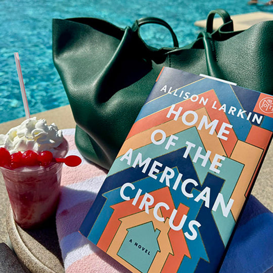 A poolside setting with a large dark green tote bag, a book and a pink drink with whipped cream and cherries