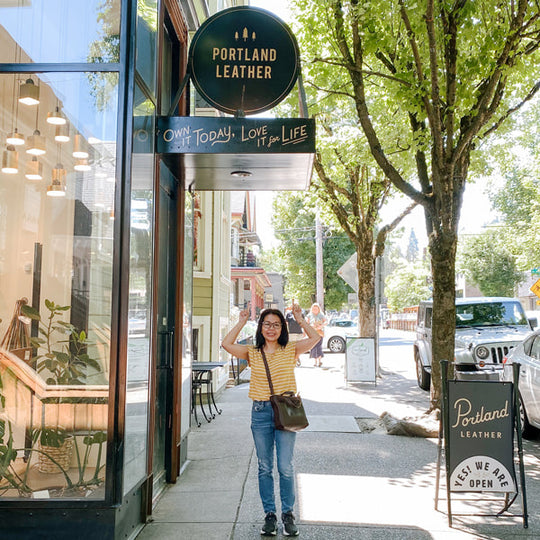 Woman standing outside 'Portland Leather' store with arms raised wearing a Mini Crossbody Tote.