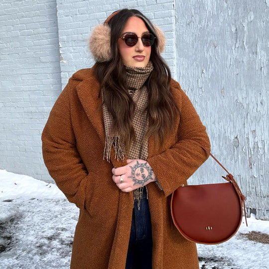 Woman in a brown coat with fur ear muffs and sunglasses, holding a brown leather bag
