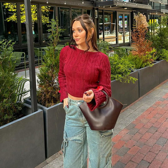 Woman in red top and blue jeans holding a brown bag on a sidewalk with plants and a building in the background