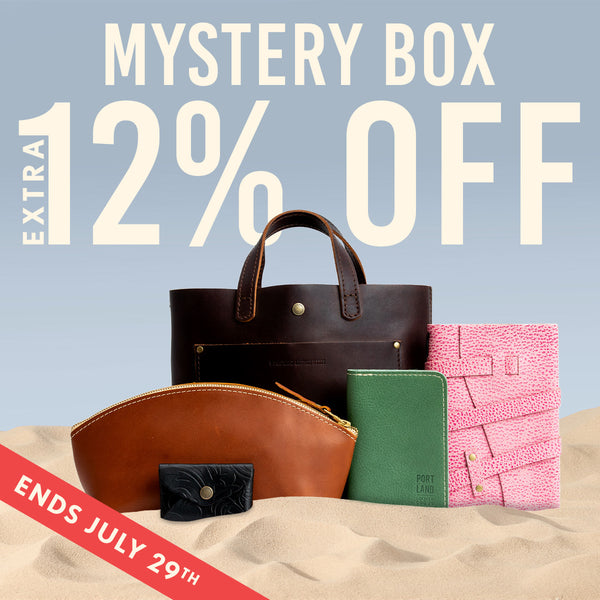Promotional graphic for a Port Lane 'Mystery Box' sale, featuring a dark brown leather tote bag, a brown leather zip pouch, a green leather notebook, a pink textured leather folio, and a small black leather cardholder arranged on a sandy surface against a light blue sky background