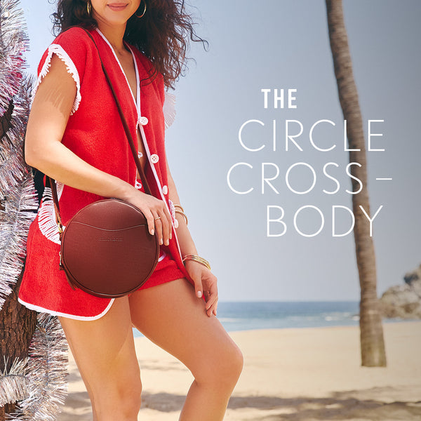A woman on a beach leaning against a tree wearing red carrying a brown tote bag