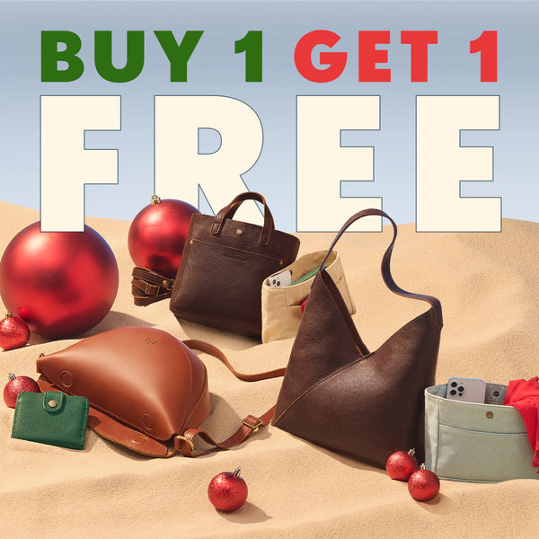 Promotional graphic with large text reading 'BUY 1 GET 1 FREE' over a styled arrangement of several handbags and small accessories on a sandy surface with red Christmas ornaments