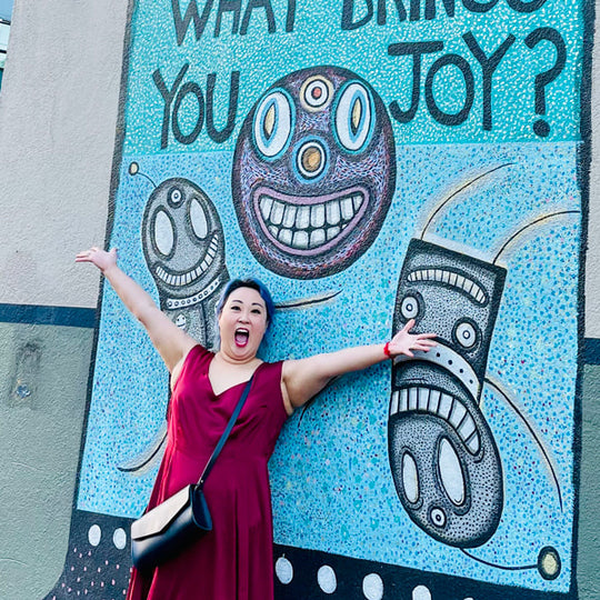 Person in a red dress wearing an archived PLG bag standing in front of a colorful mural with question 'What Brings You Joy?'