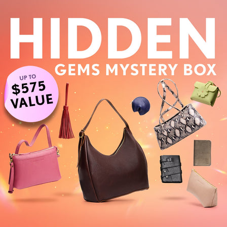 Promotional graphic for a 'hidden Gems Mystery Box' featuring a collection of shoulder bags, crossbodies, wallets, and small goods. Set against a orange sparkly background with a pink badge indicating up to $575 value.