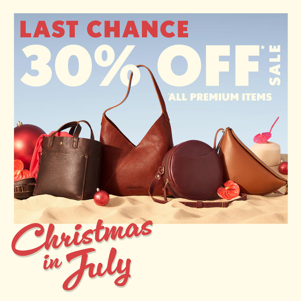 Promotional sale graphic for a July 30th Christmas sale, featuring a collection of brown and burgundy handbags on a sandy beach with Christmas ornaments under a light blue sky. Text reads 'LAST CHANCE 30% OFF SALE' and 'Christmas in July'