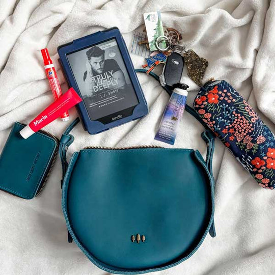 Moon shaped leather bag surrounded by its contents such as wallet, kindle, glasses case, and keys