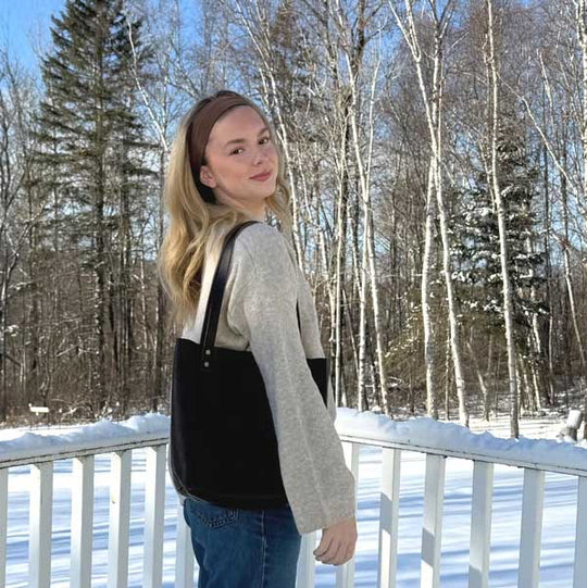 Woman wearing headband and carrying a dark leather tote bag standing in front of wintery woods scenery