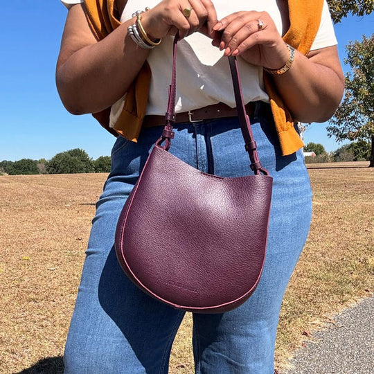 Person holding a Plum Olivia Shoulder Bag outdoors with a clear sky and trees in the background