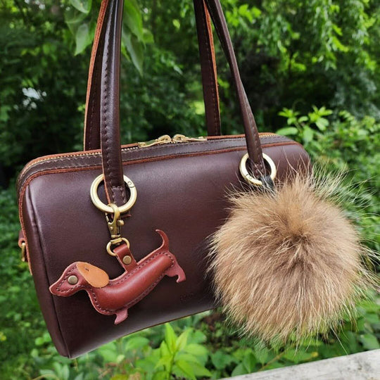 Sloan Doctor Bag with a Doxie Keychain and fur pom-pom against a green foliage background