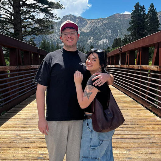 Two people standing on a wooden bridge with mountains in the background with a Flora Shoulder Bag