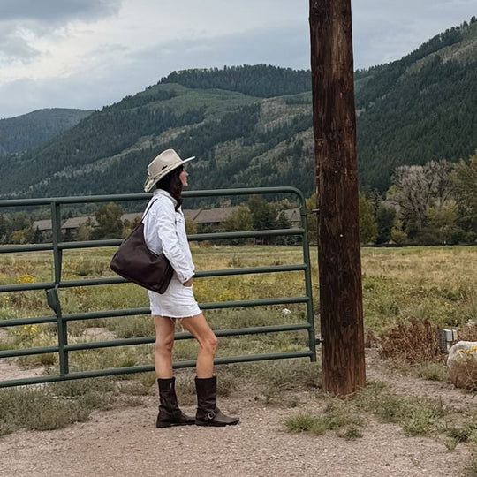 Person standing by a gate in a scenic mountainous area wearing an Aspen Shoulder Bag