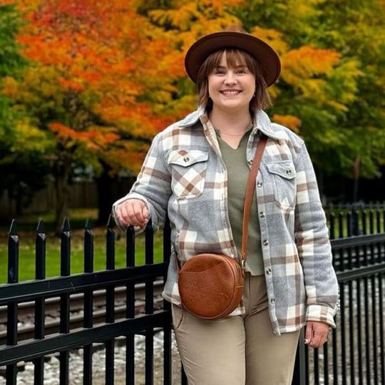 Person wearing a plaid shirt and brown hat with a Circle Crossbody, standing in front of autumn trees.