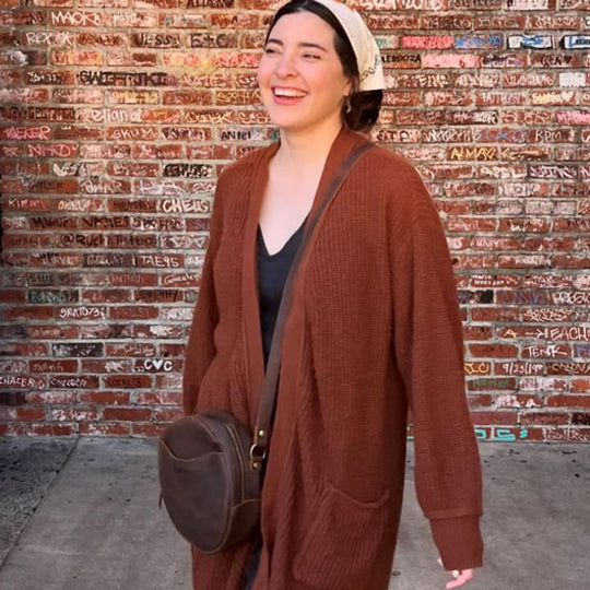 Woman wearing a brown cardigan and carrying a Circle Crossbody against a brick wall.