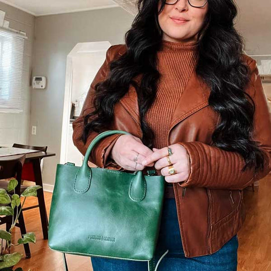 Woman holding a green handbag indoors, wearing a brown leather jacket and turtleneck.