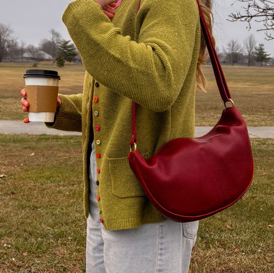 Person wearing a green sweater and holding a Chili Red Koala Sling Bag and coffee cup outdoors.