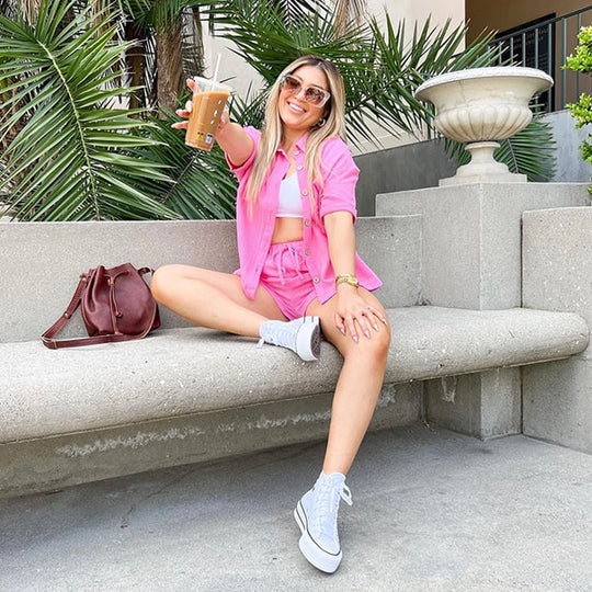 Women sitting on a concert bench with a brown bucket bag, wearing a pink outfit holding out a coffee
