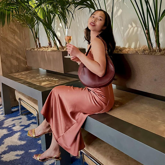 Women wearing a brown bag on her shoulder sitting on a bench with a drink in her hand