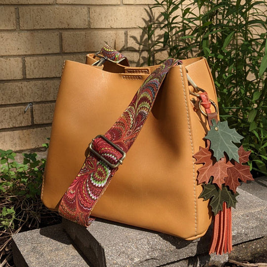 Mango Butterfly Bucket Bag with a colorful strap and a maple leaf tassel resting on stone