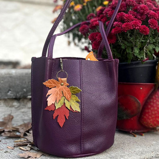 A plum Devan Bucket Tote with a maple leaf tassel sitting in front of mums in a pot