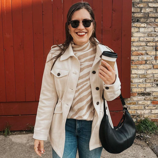 A person standing in front of a fence holding a hot drink and carrying the Nora Shoulder Bag in Black