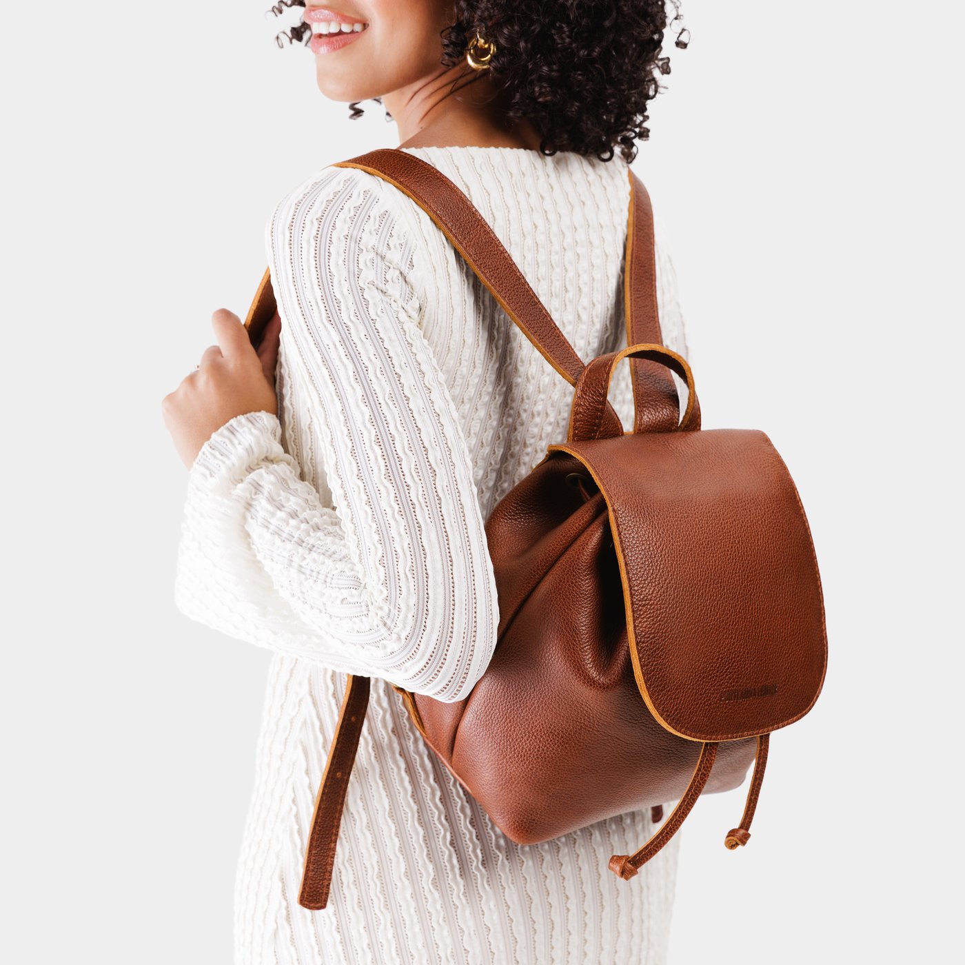 Nutmeg | Pebbled brown leather drawstring backpack being worn by a person wearing a white dress.