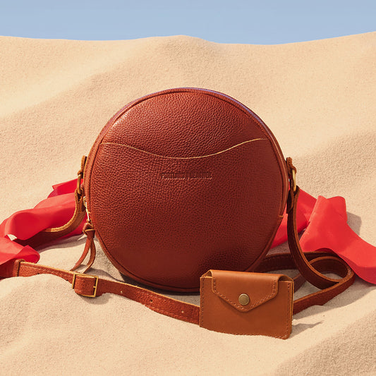 A round brown pebbled leather crossbody bag with a matching small pouch and strap, styled on sand 