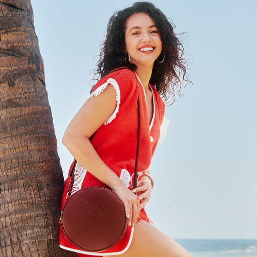 A woman on a beach leaning against a tree wearing red carrying a brown tote bag featured image