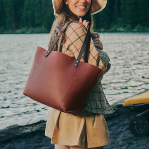 A woman outdoors by the water wearing a wide-brim hat and carrying a large brown leather tote bag over her shoulder.