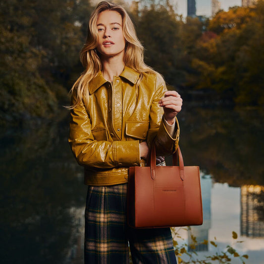 A woman standing outdoors by a reflective lake at dusk, wearing a glossy mustard yellow leather jacket and plaid pants, while holding a  Honey Lola Tote Bag.