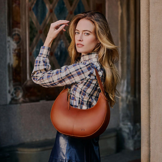 A woman wearing a blue and beige plaid shirt and a dark skirt carries a Honey Nora Shoulder Bag, set against a blurred architectural background.