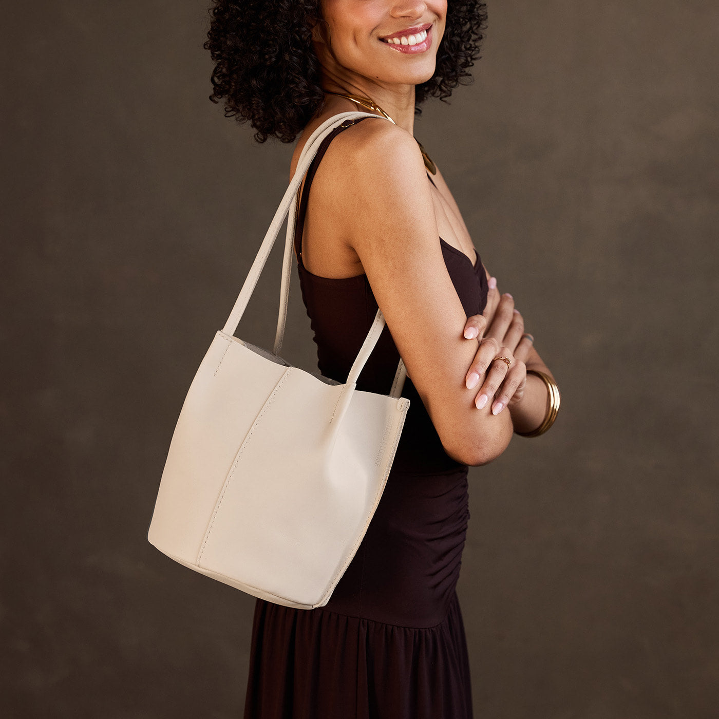 Bone*Classic | Model wearing sleeveless dark brown dress carrying petite bucket shaped tote bag with matching leather handles against dark grey background