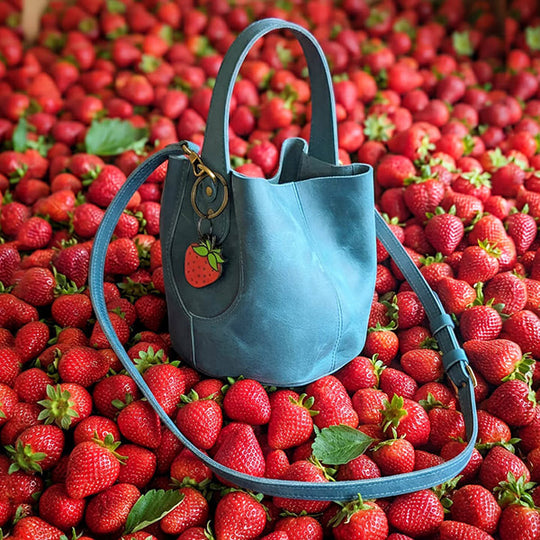 A blue leather handbag with a strawberry charm, placed on a bed of fresh red strawberries