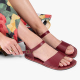 Marsala | Close-up of a person wearing colorful dress and red flat sandals with an ankle strap and buckle, shown against a plain background.