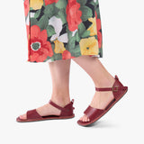 Marsala | Close-up of a person wearing colorful dress and red flat sandals with an ankle strap and buckle, shown against a plain background.