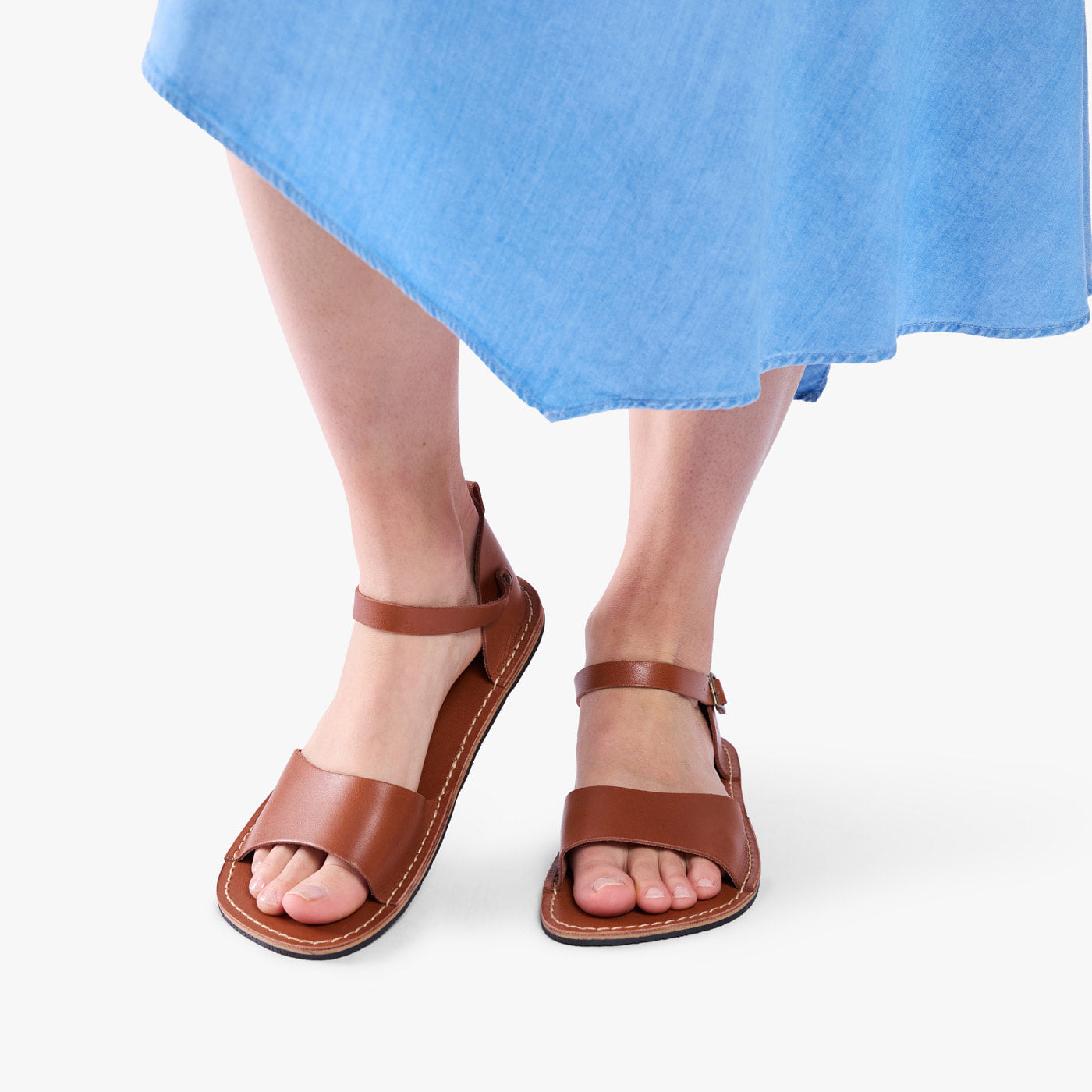 Miele | Person wearing blue dress and tan flat sandals with an ankle strap and buckle, shown against a plain background.