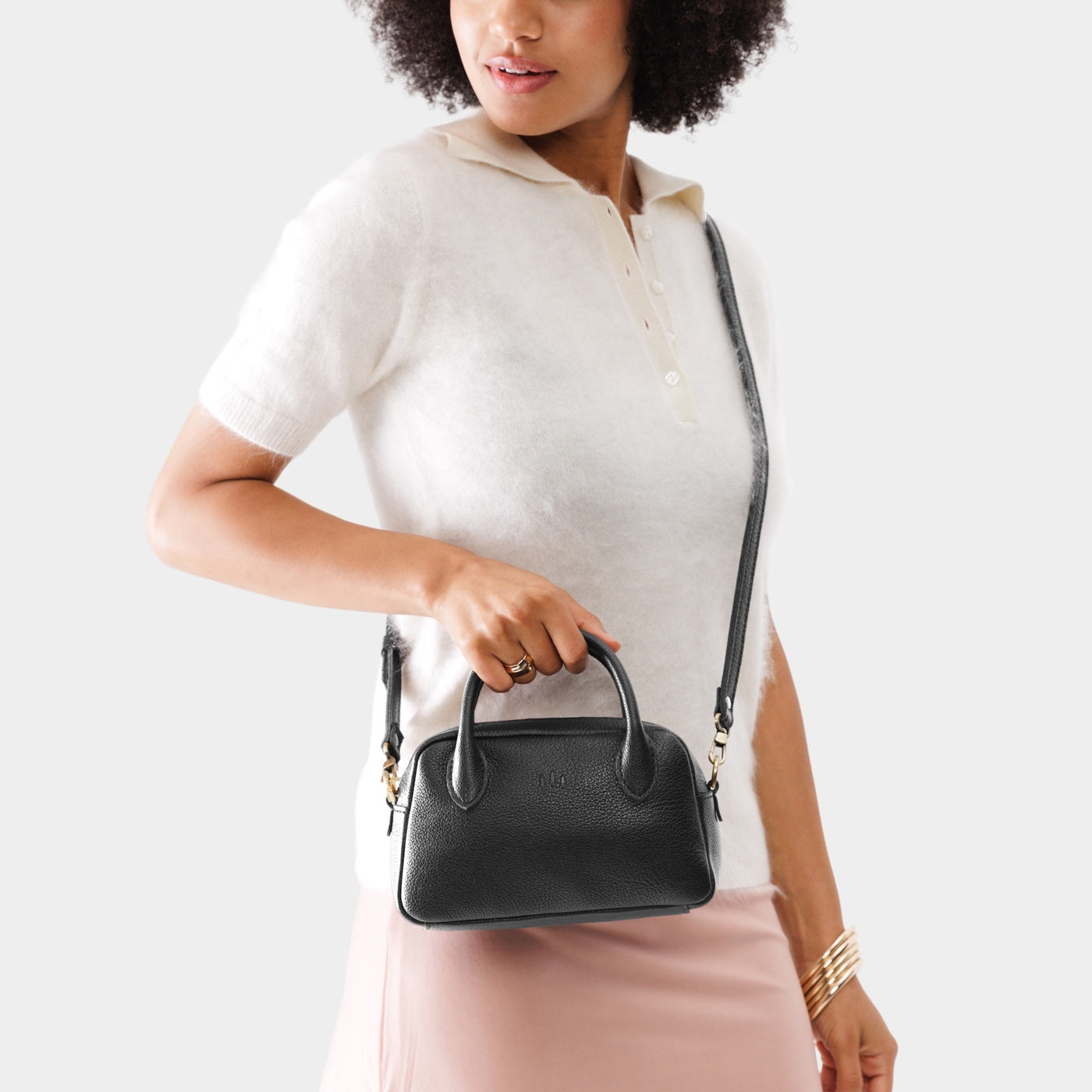 Pebbled--black*Small | A bowler styled bag with a crossbody strap being held by a model wearing a dress