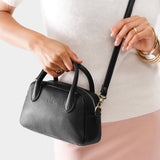 Pebbled--black Small | A bowler styled bag with a crossbody strap being held by a model wearing a dress