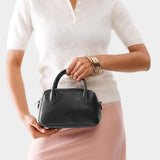 Pebbled--black Small | A bowler styled bag with a crossbody strap being held by a model wearing a dress