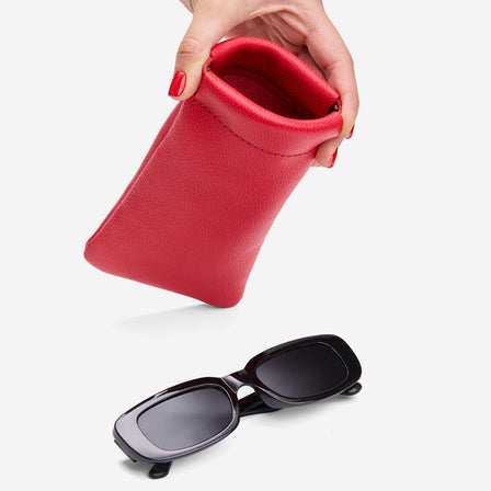 Cherry | Model hand holding open leather glasses case with hidden metal spring closure above a pair of sunglasses