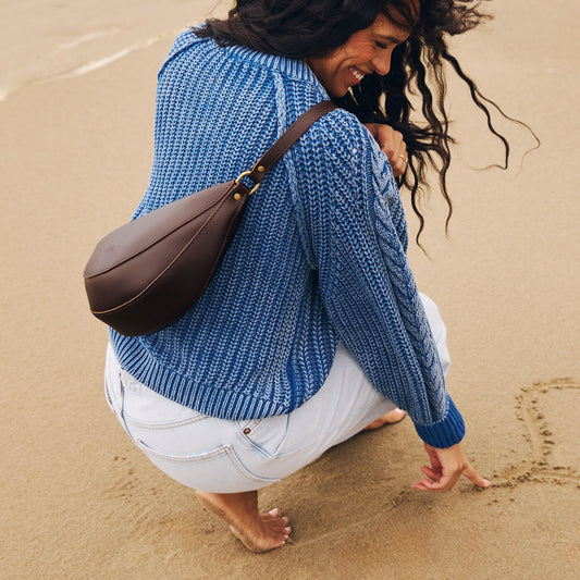 A model crouching on a sandy beach wearing a Grizzly Koala Sling Bag.
