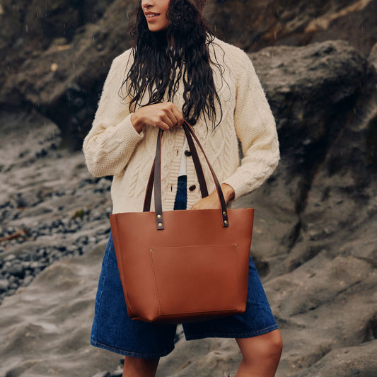 Model wearing knitted button up cardigan carrying a leather tote bag in honey against a pnw beach background