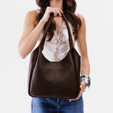 Coldbrew Large | A model holding a u-shaped, flat-bottomed leather shoulder bag with two outside pockets