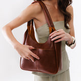 Nutmeg Large | A model holding a u-shaped, flat-bottomed leather shoulder bag with two outside pockets