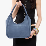 Denim Medium | A model holding a u-shaped, flat-bottomed leather shoulder bag with two outside pockets