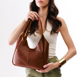 Nutmeg Medium | A model holding a u-shaped, flat-bottomed leather shoulder bag with two outside pockets