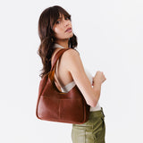 Nutmeg Medium | A model holding a u-shaped, flat-bottomed leather shoulder bag with two outside pockets