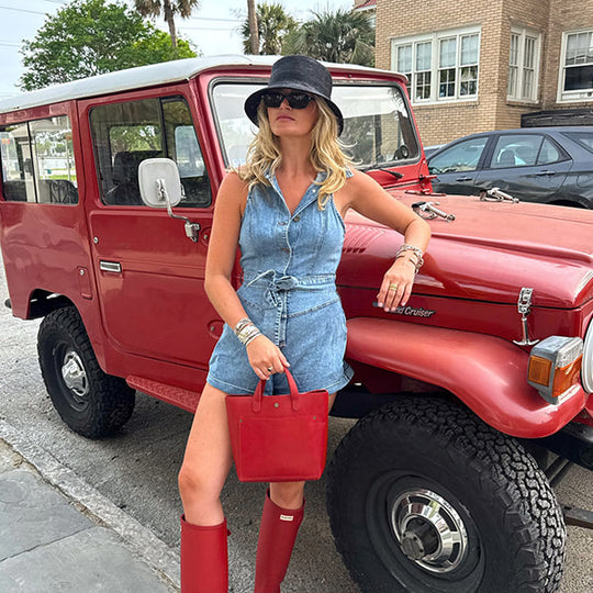 A woman posing outdoors in front of a red vintage jeep holding a red handbag